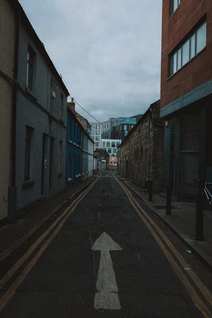 A narrow residential street featuring a row of colorful terraced houses with white, yellow, and blue facades, situated on the right side of the image. The street is paved with asphalt and has a pair o