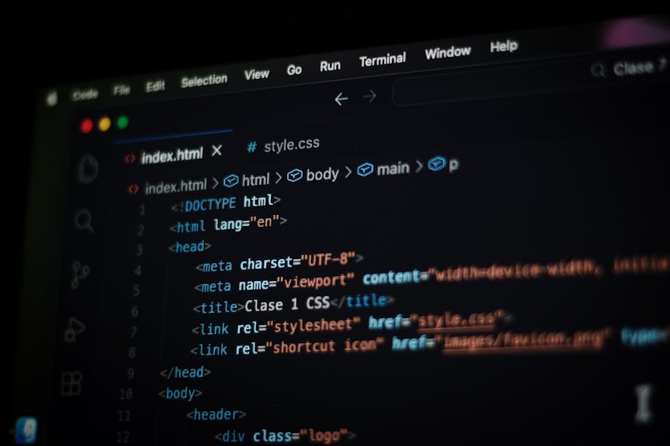 A person with dark hair, seen from behind, working on a laptop displaying a screen filled with colorful lines of computer code. The workspace includes a large desktop monitor, a notebook, and a smartp